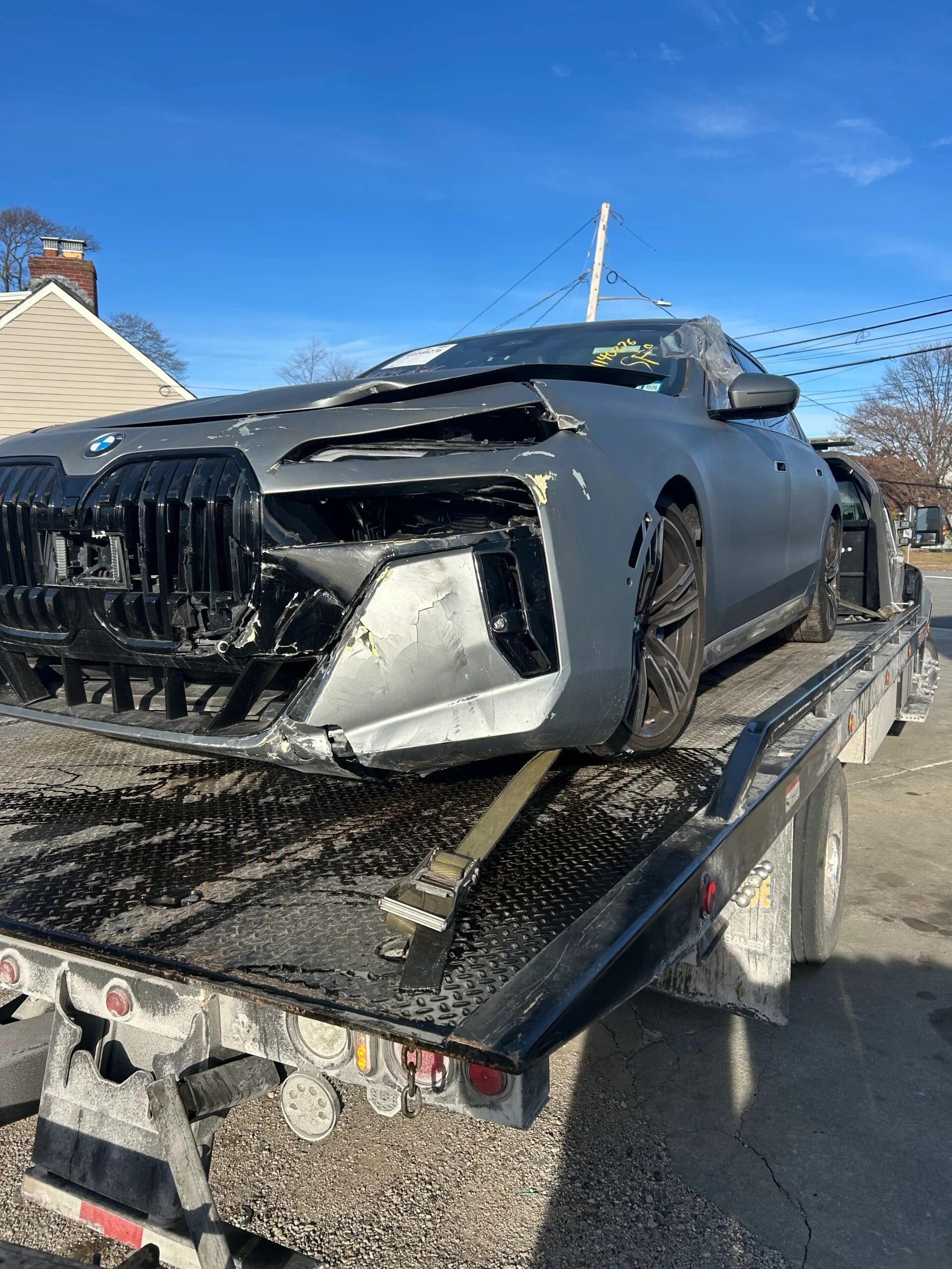 Damaged black sports car on a tow truck, front end heavily smashed in a snowy neighborhood.