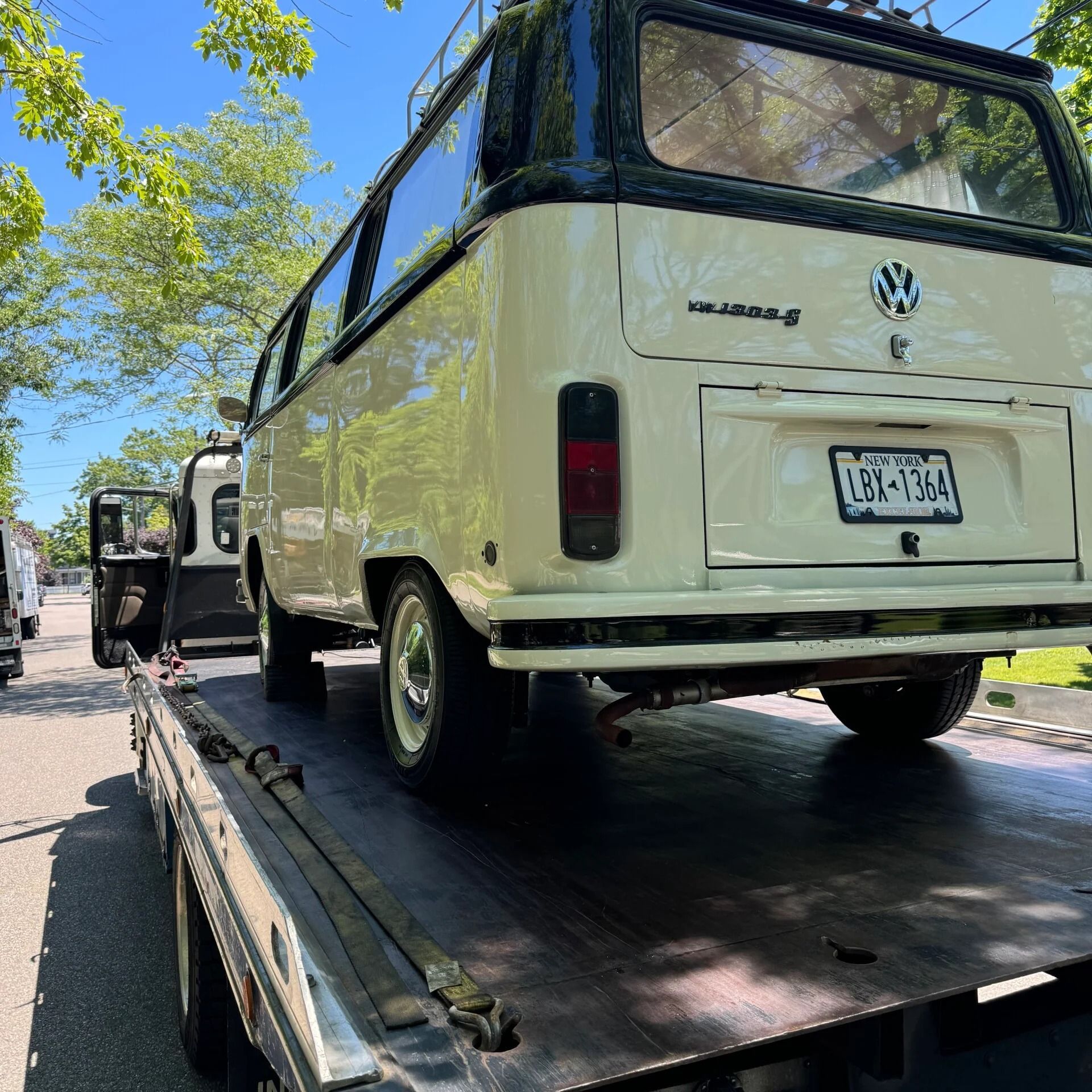 Cream-colored VW van on a flatbed tow truck under trees on a sunny street.