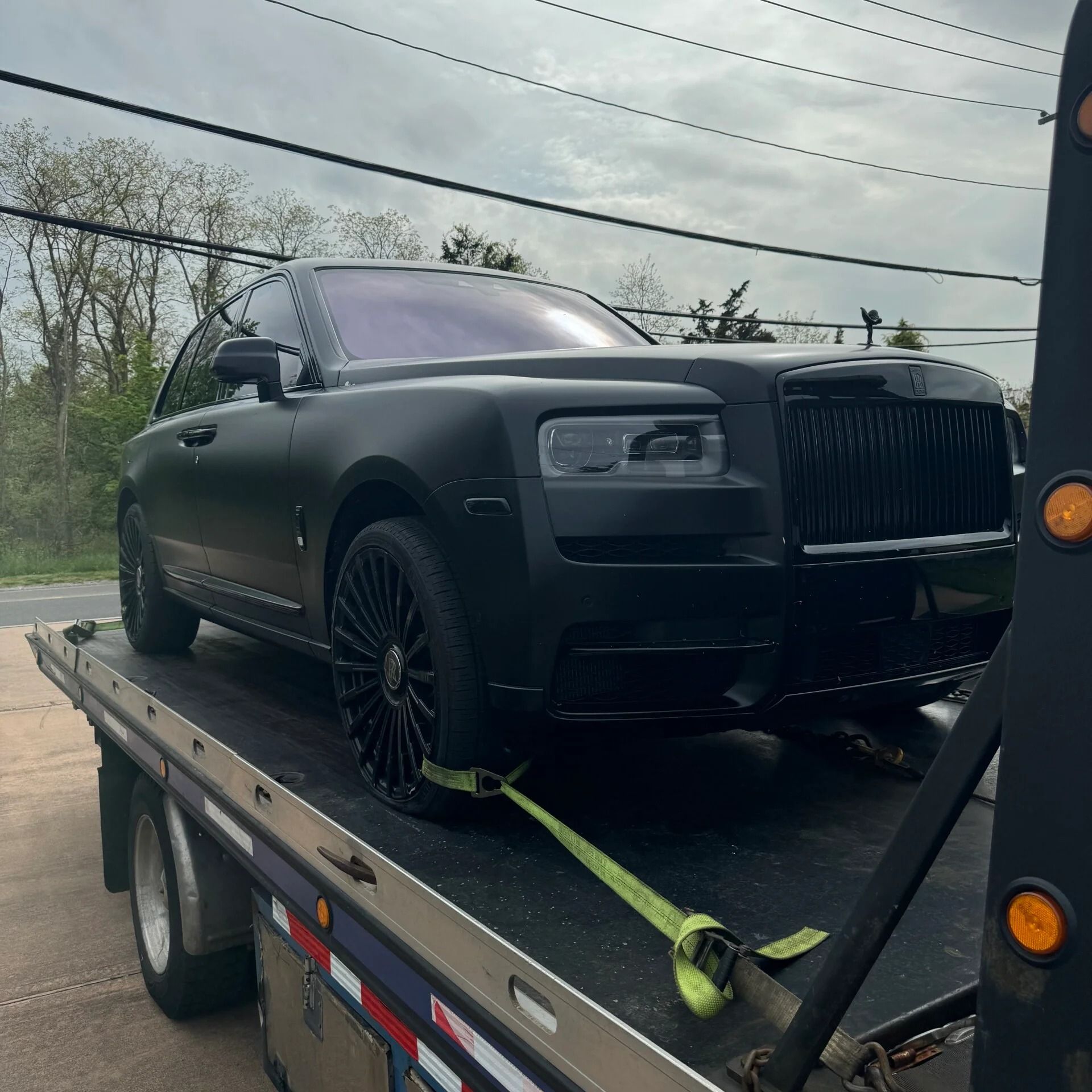 Black luxury SUV strapped on a flatbed tow truck in a rainy outdoor lot