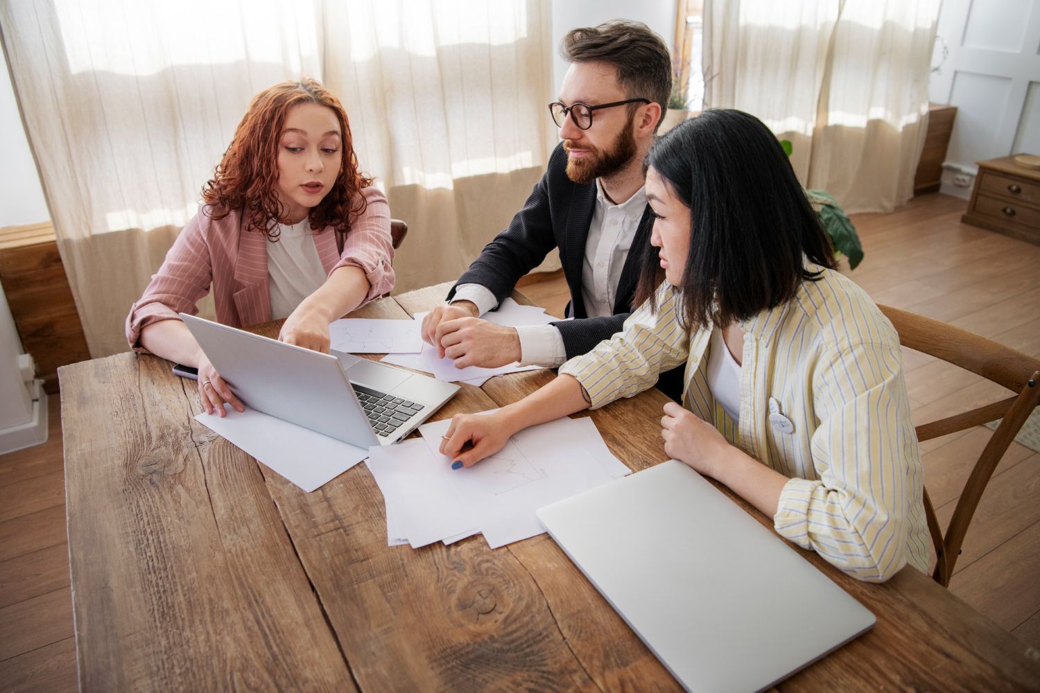 Three coworkers discussing papers and a laptop at a wooden table in a bright office.