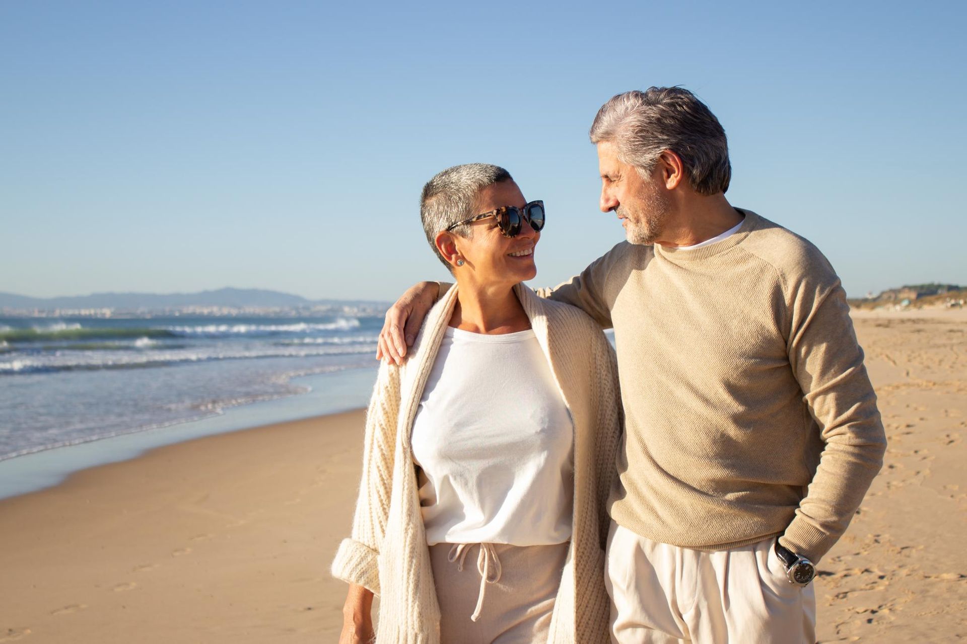 Couple walking arm in arm on a sunny beach, smiling at each other by the ocean.