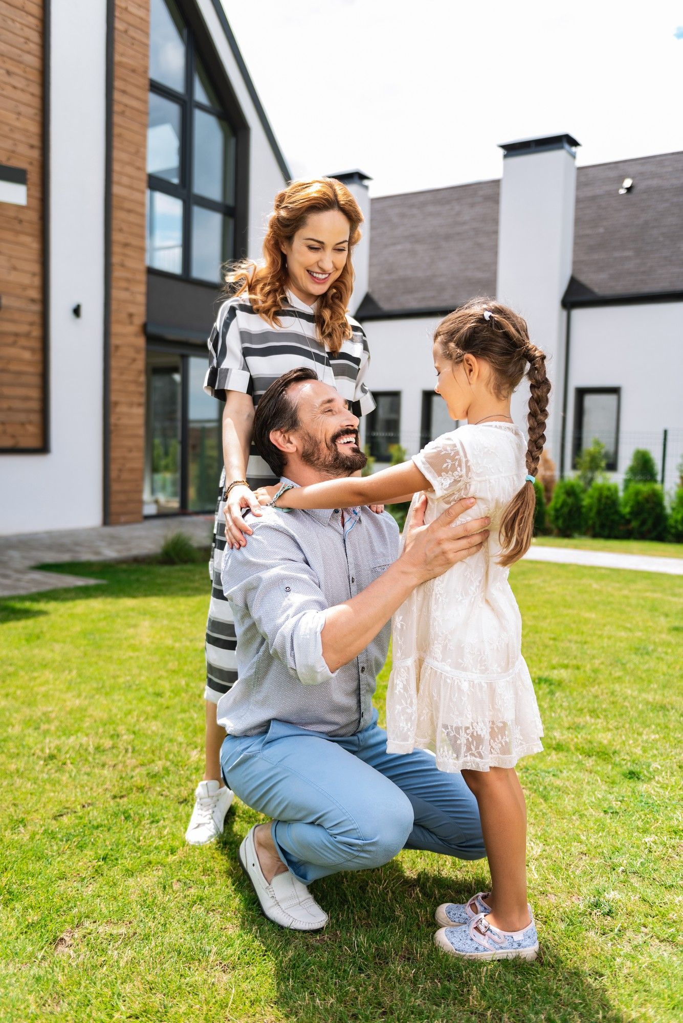 Man kneeling on lawn hugging a young girl while a woman stands beside them outside a modern house