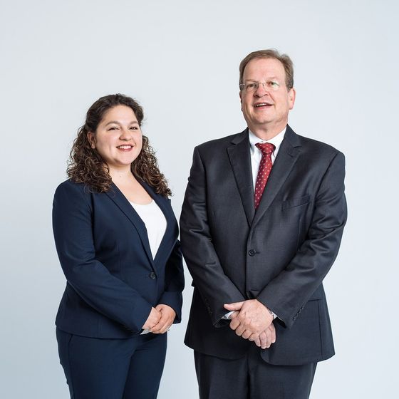 Two business professionals in dark suits standing and smiling against a light gray background
