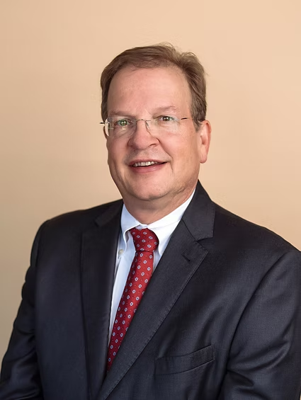 Portrait of a smiling man in a dark suit, white shirt, and red tie against a beige background