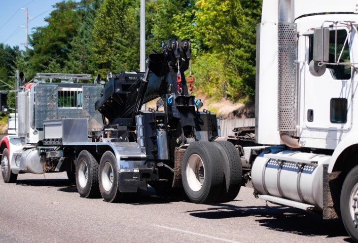 Tow truck hauling a damaged black vehicle on a street, with another truck beside it