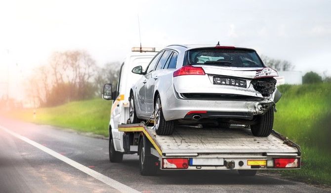 Silver car on a tow truck beside a road in a grassy area.