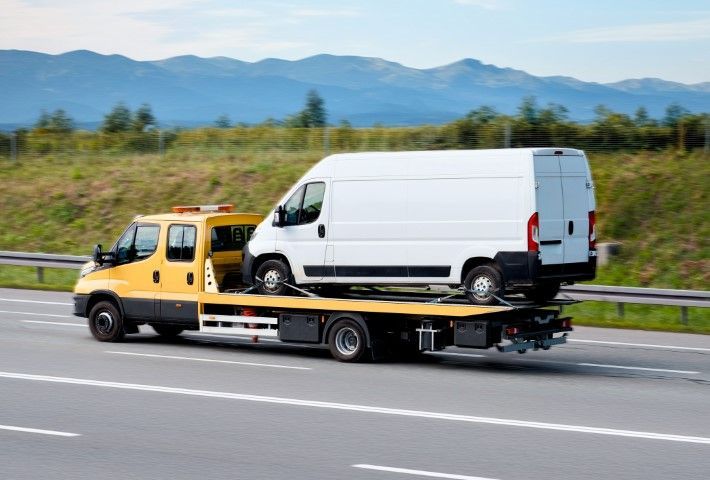 Yellow tow truck carrying a white cargo van on a highway with mountains in the background