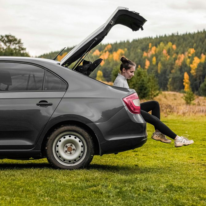 Person sitting on the open trunk of a gray car in a grassy field with autumn trees in the background