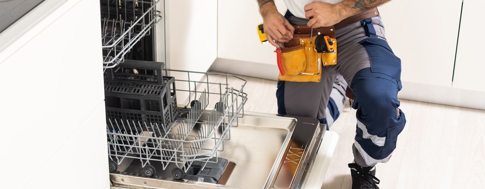 Person in workwear with a tool belt, working on an open dishwasher in a kitchen.