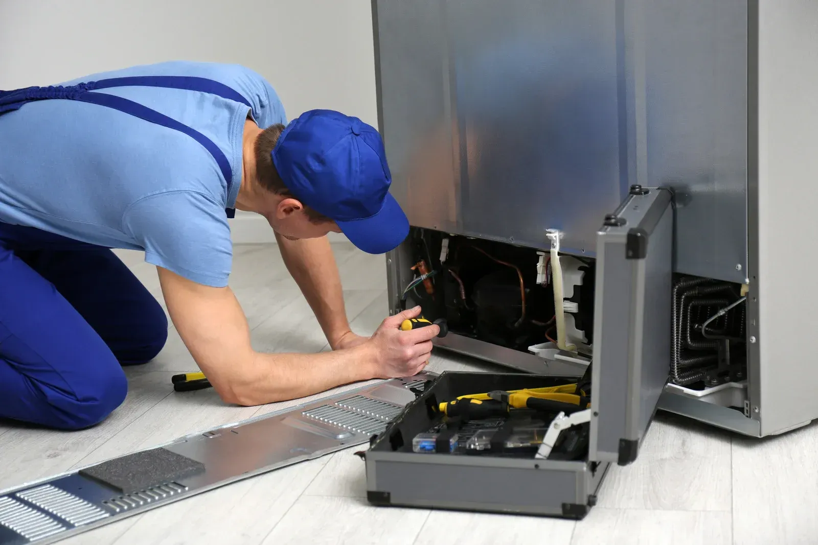 A person in blue jumpsuit repairs a refrigerator, using tools from an open toolbox on the floor.