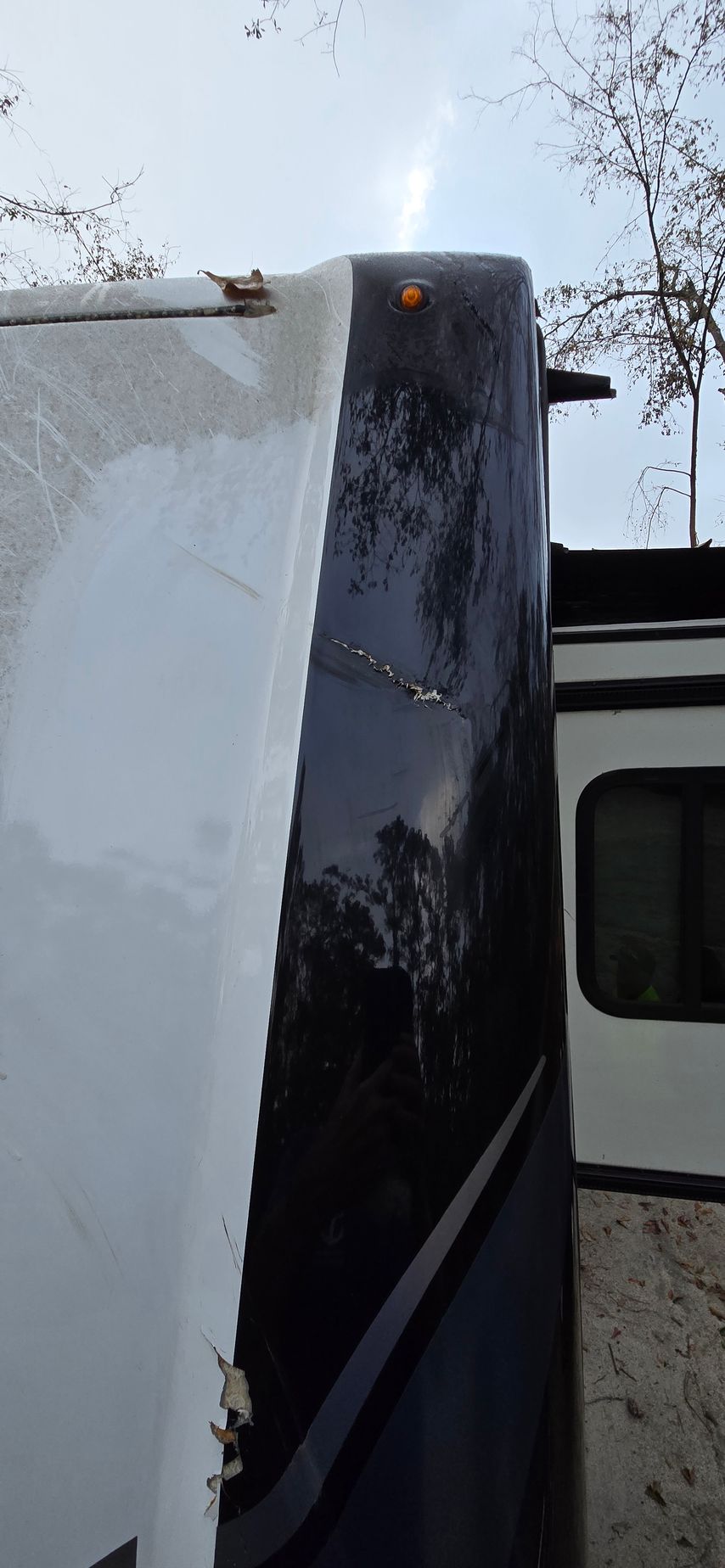 Close-up of a damaged, peeling black front cap on an RV exterior with a white side panel and trees in the background.