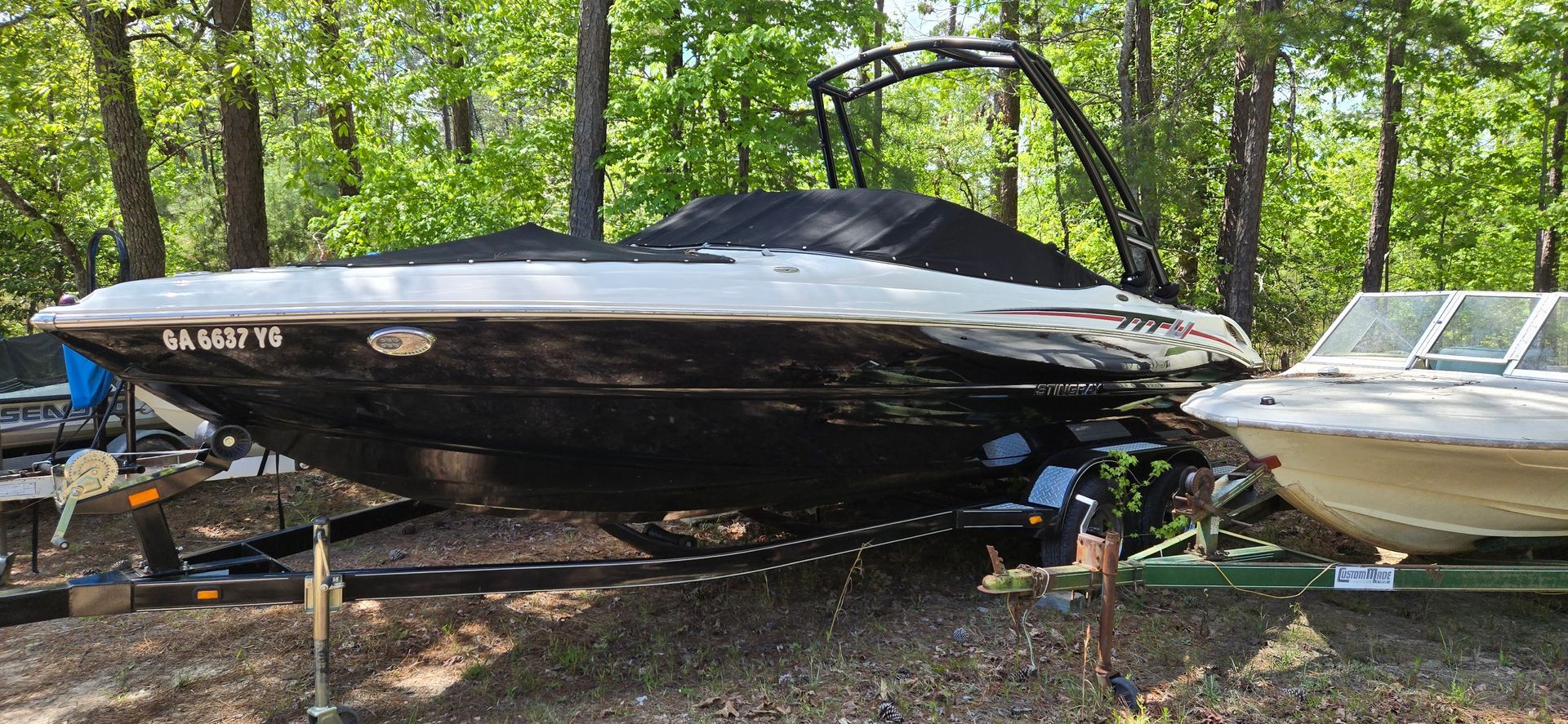 A black and white motorboat with a tower, covered by a black tarp, sits on a trailer in a wooded outdoor boat storage lot.