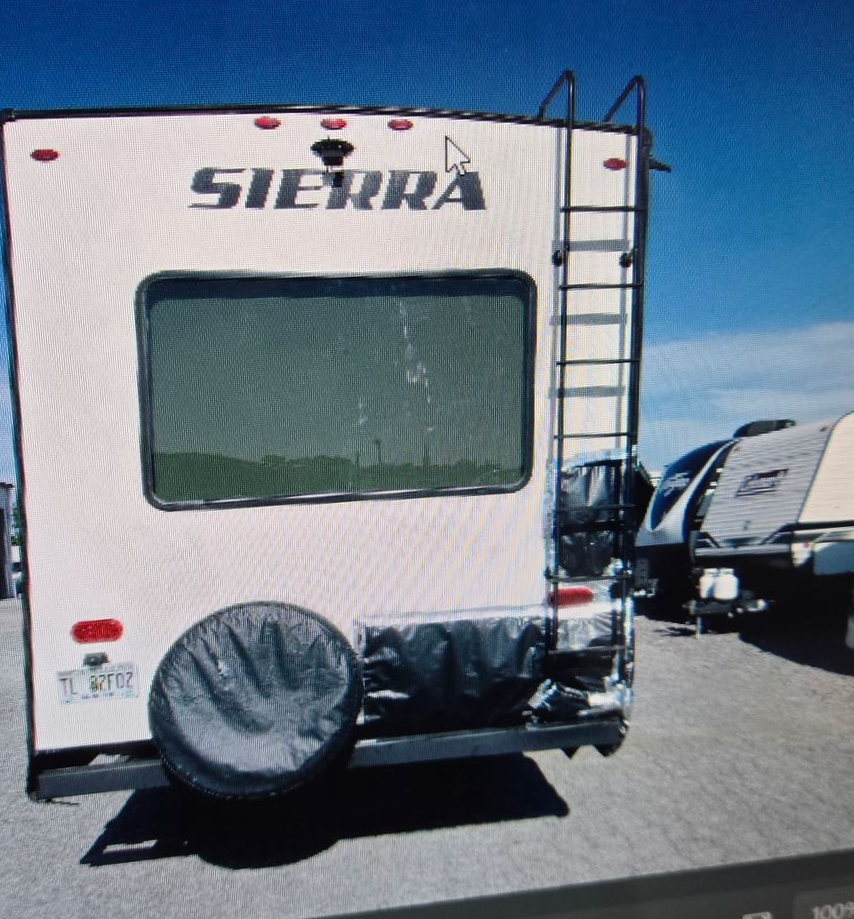 The rear view of a white Sierra RV with a black ladder, spare tire cover, and bumper, parked on a gravel lot.