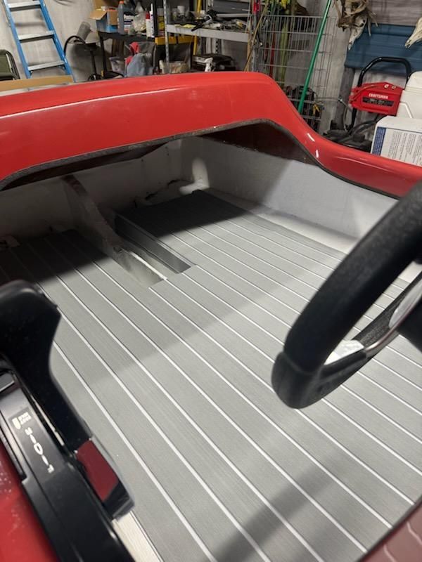 The interior of a red boat features gray faux-teak flooring with white stripes, a steering wheel, and a throttle lever.