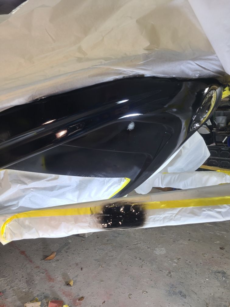 A close-up of a glossy black car part being masked with white plastic and yellow tape for a paint job.
