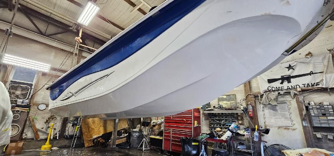 A white and blue motorboat hangs suspended in a well-lit workshop with tools and equipment in the background.