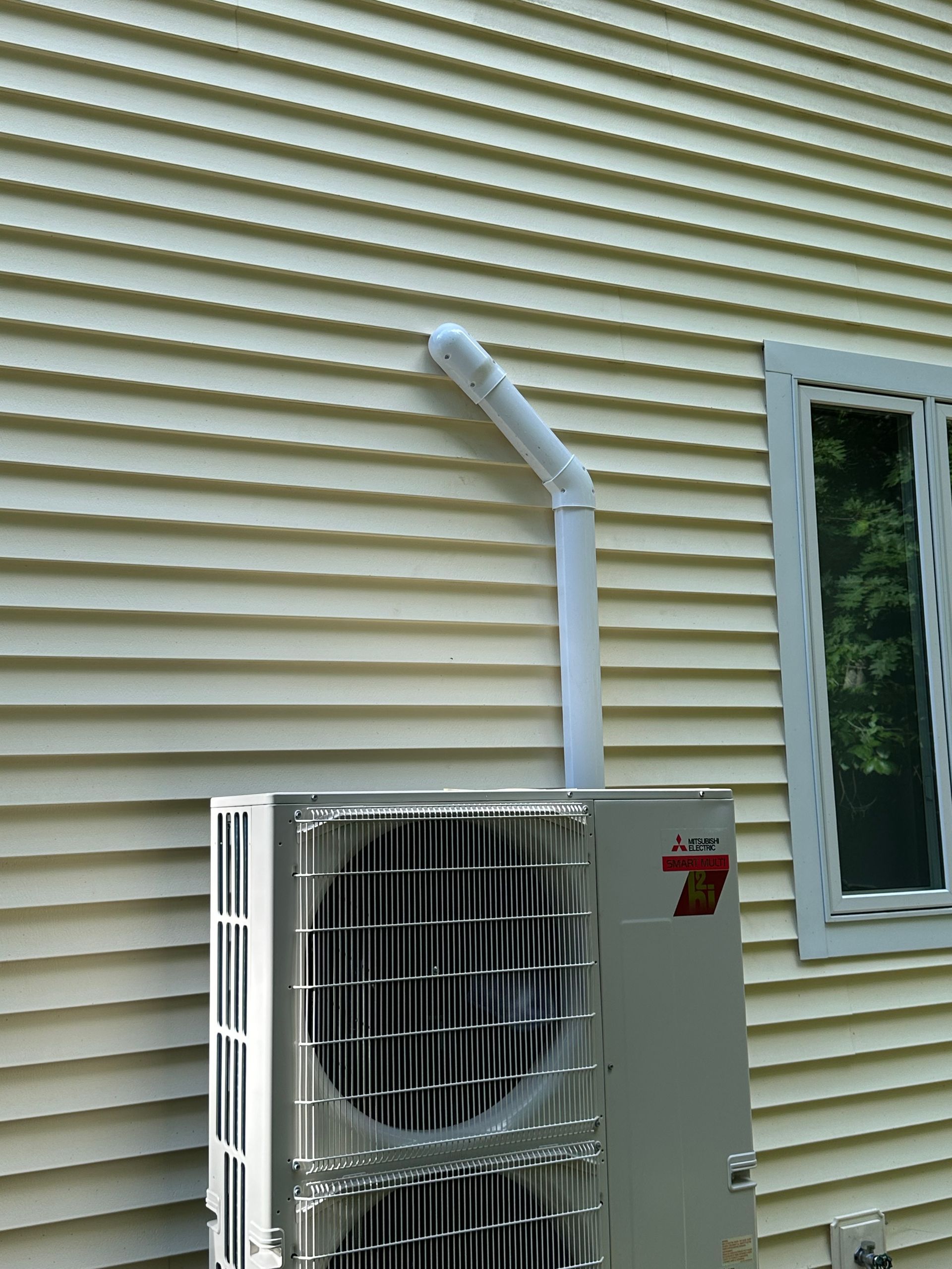 An outdoor HVAC heat pump unit sits against light yellow horizontal siding, connected to the wall by a white pipe.