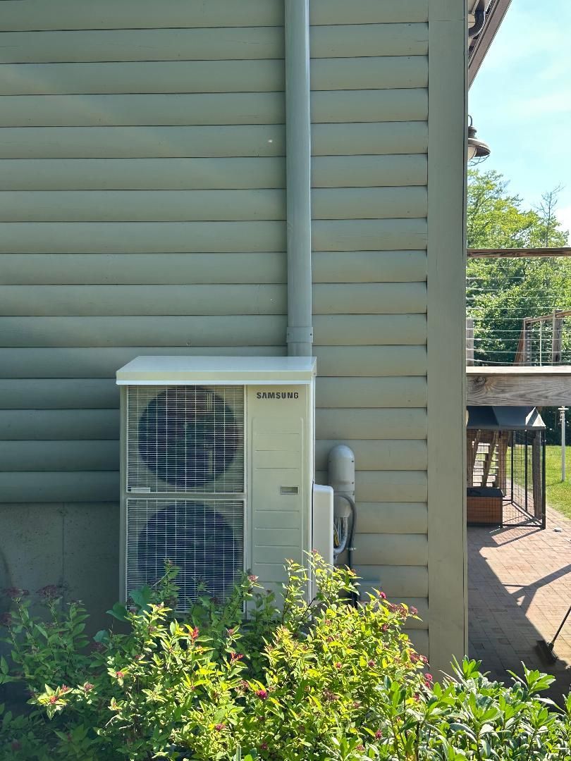 A light gray outdoor HVAC unit sits against the side of a house with green horizontal siding, partially hidden by bushes.