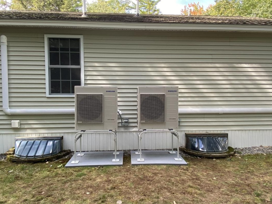 Two outdoor HVAC units on metal stands against a beige house wall, with vents below the windows.