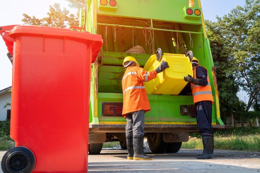 Garbage collectors emptying a yellow bin into a green truck beside a red bin on a street