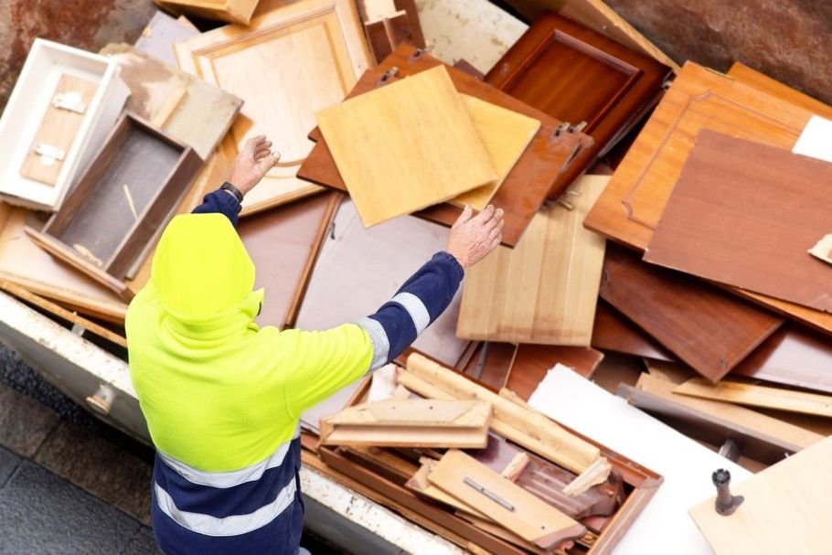 Person in a neon hoodie sorting wooden furniture pieces in a dumpster full of discarded cabinets and boards
