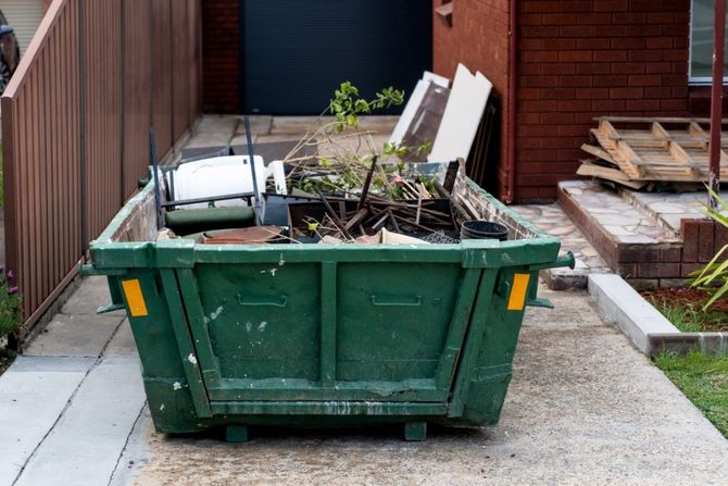Green dumpster filled with debris in a driveway beside a house