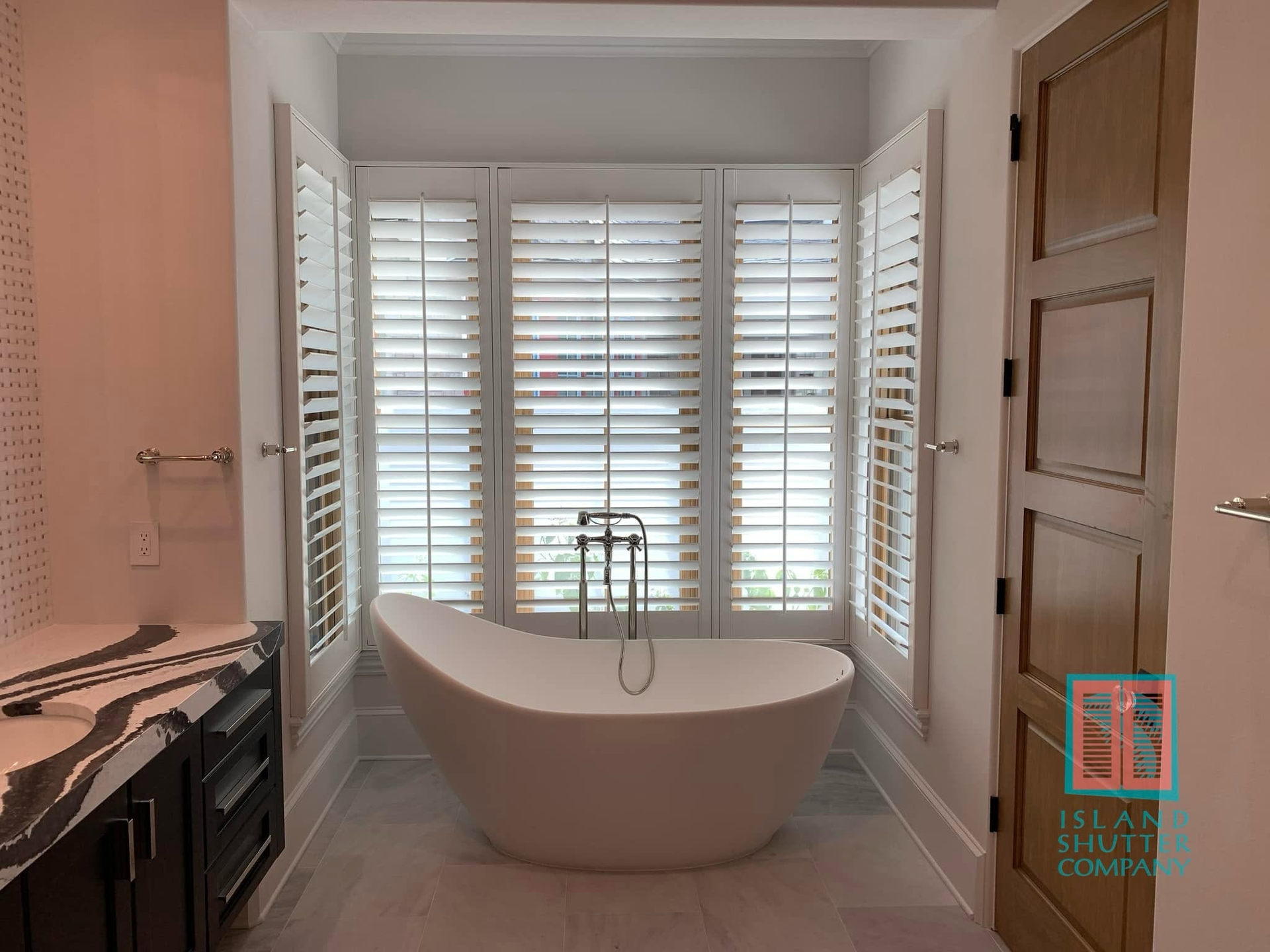 Bright bathroom with a freestanding tub, white shutters, dark vanity, and wooden door