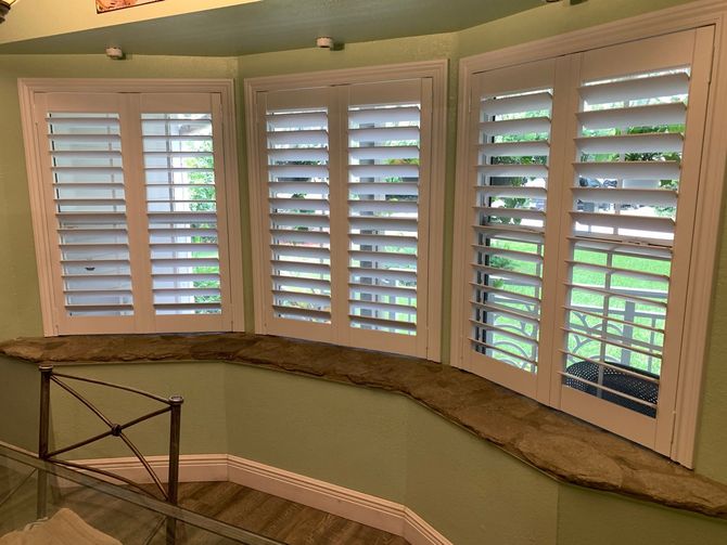 Curved bay window with white shutters, overlooking a green yard and beige walls inside a bright room