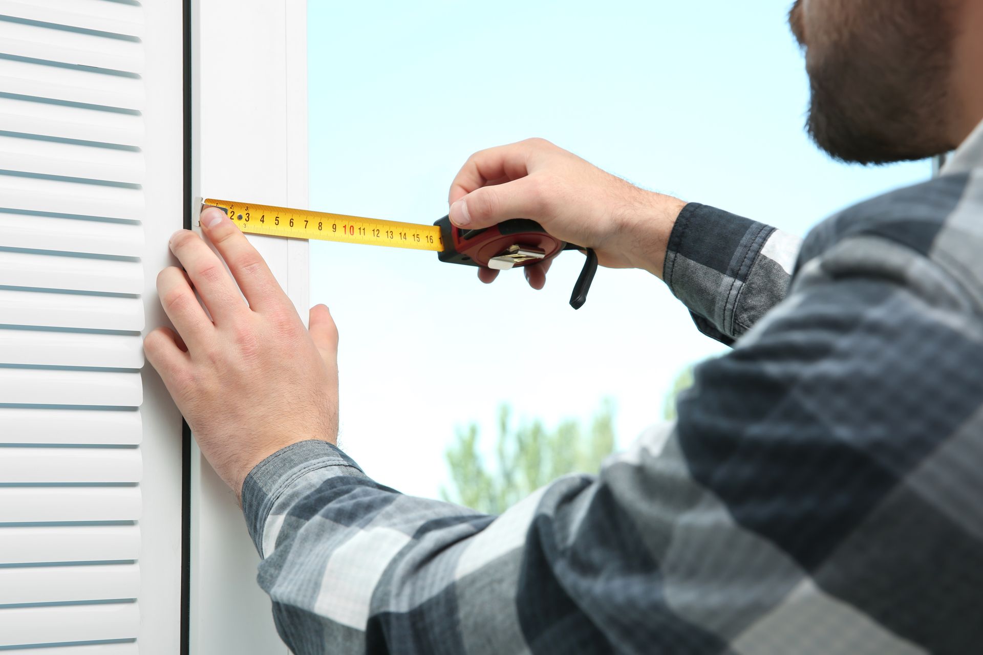 A man outdoors measuring a window with a measuring tape.