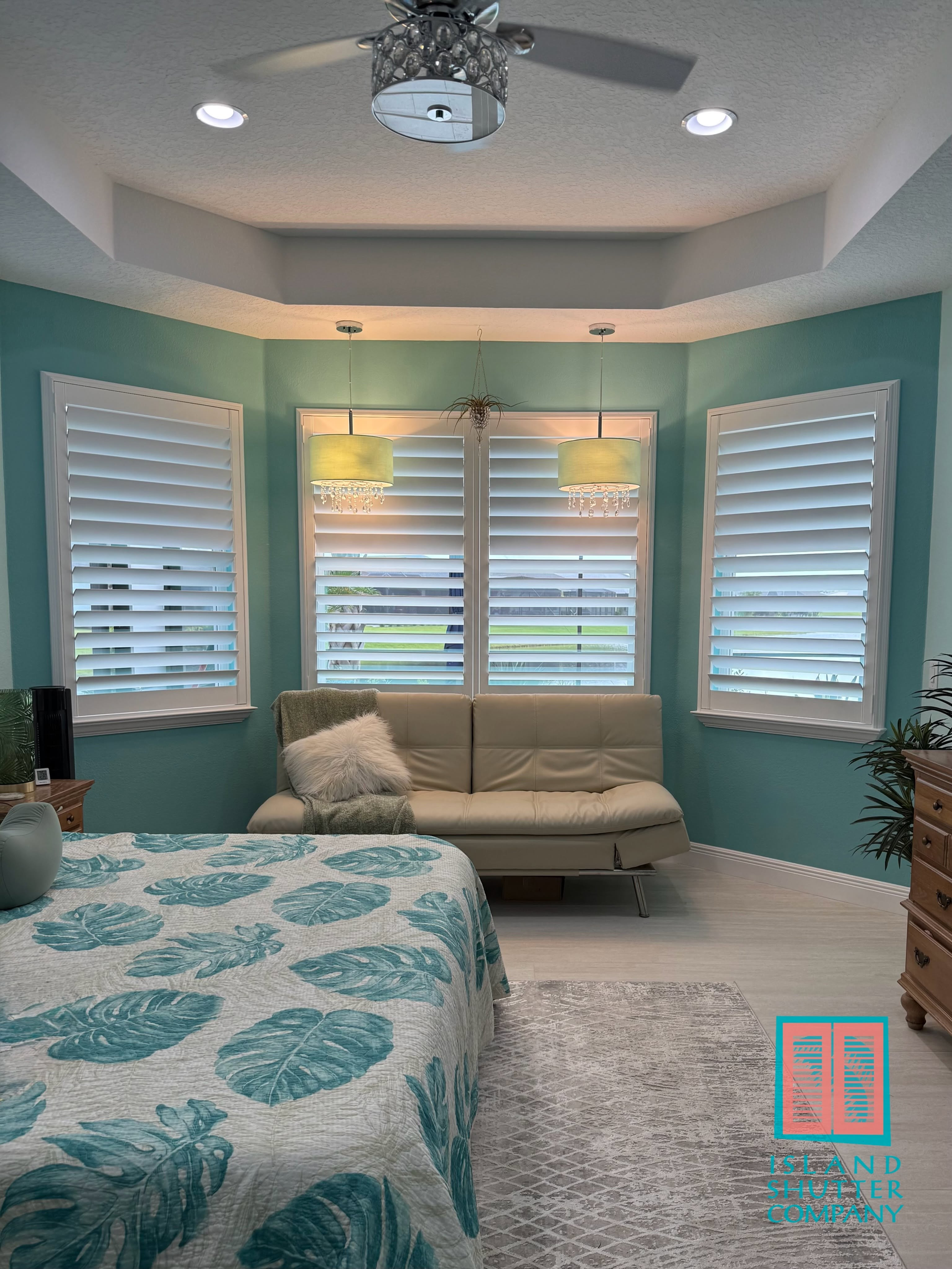Teal bedroom with bay windows, white blinds, a chaise lounge, and a ceiling fan.