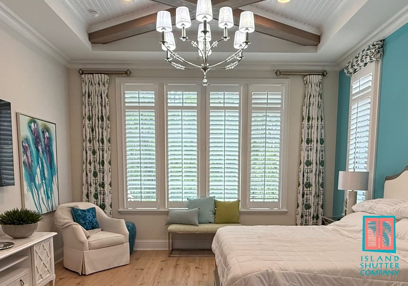 Bright bedroom with vaulted ceiling, chandelier, large windows, white bed, and teal accents