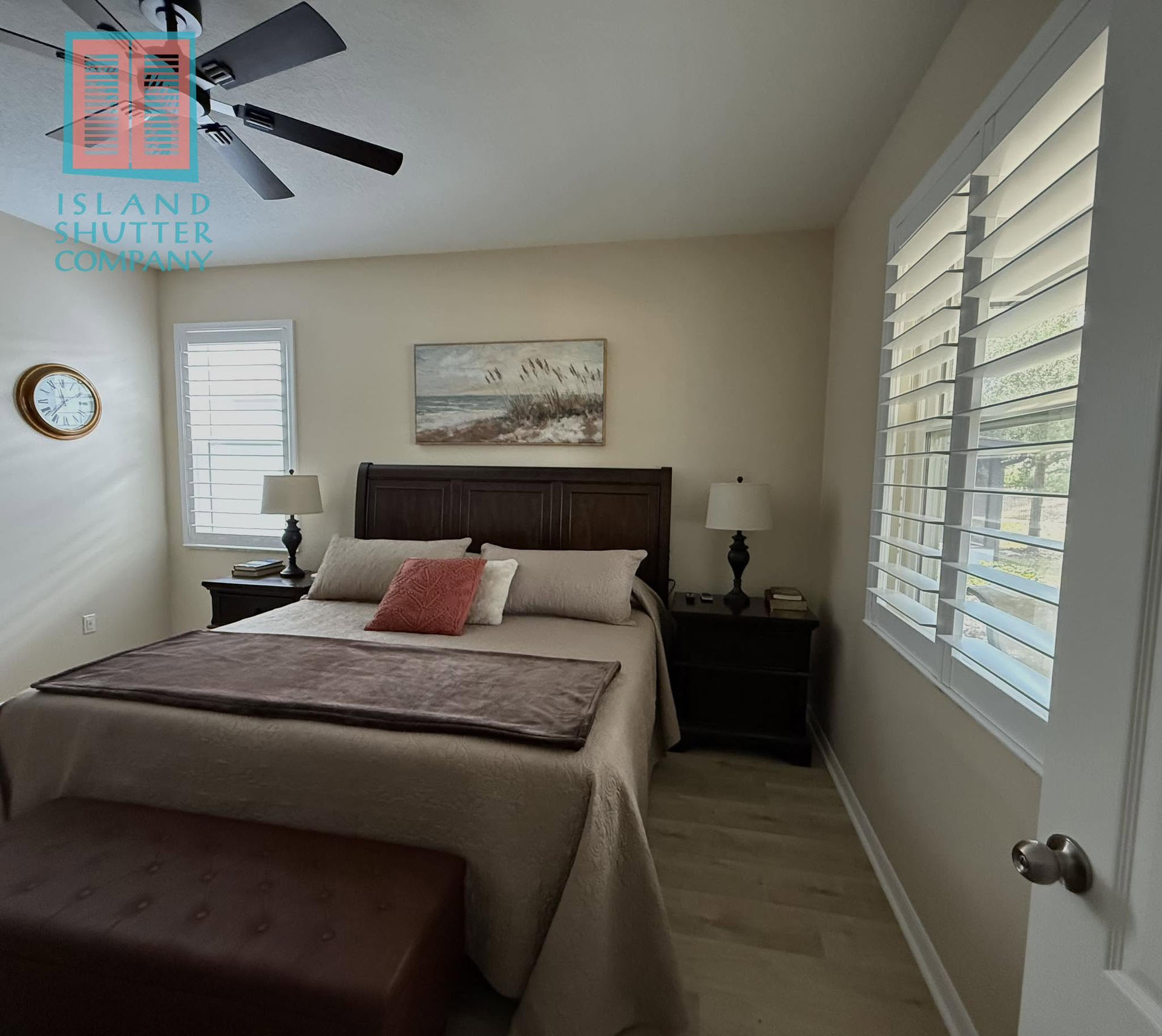 Bright bedroom with a bed, nightstands, ceiling fan, and large windows with white shutters.
