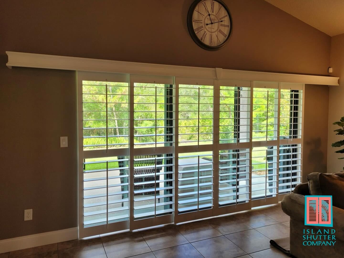 Empty living room with large windows, white blinds, wall clock, and hardwood floors