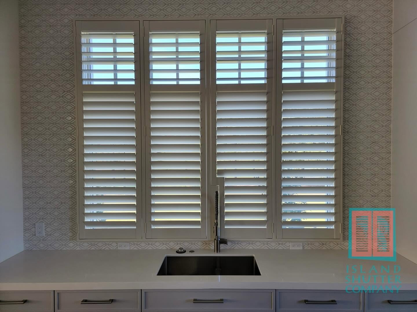 Kitchen sink beneath three shuttered windows with patterned wallpaper and white cabinets