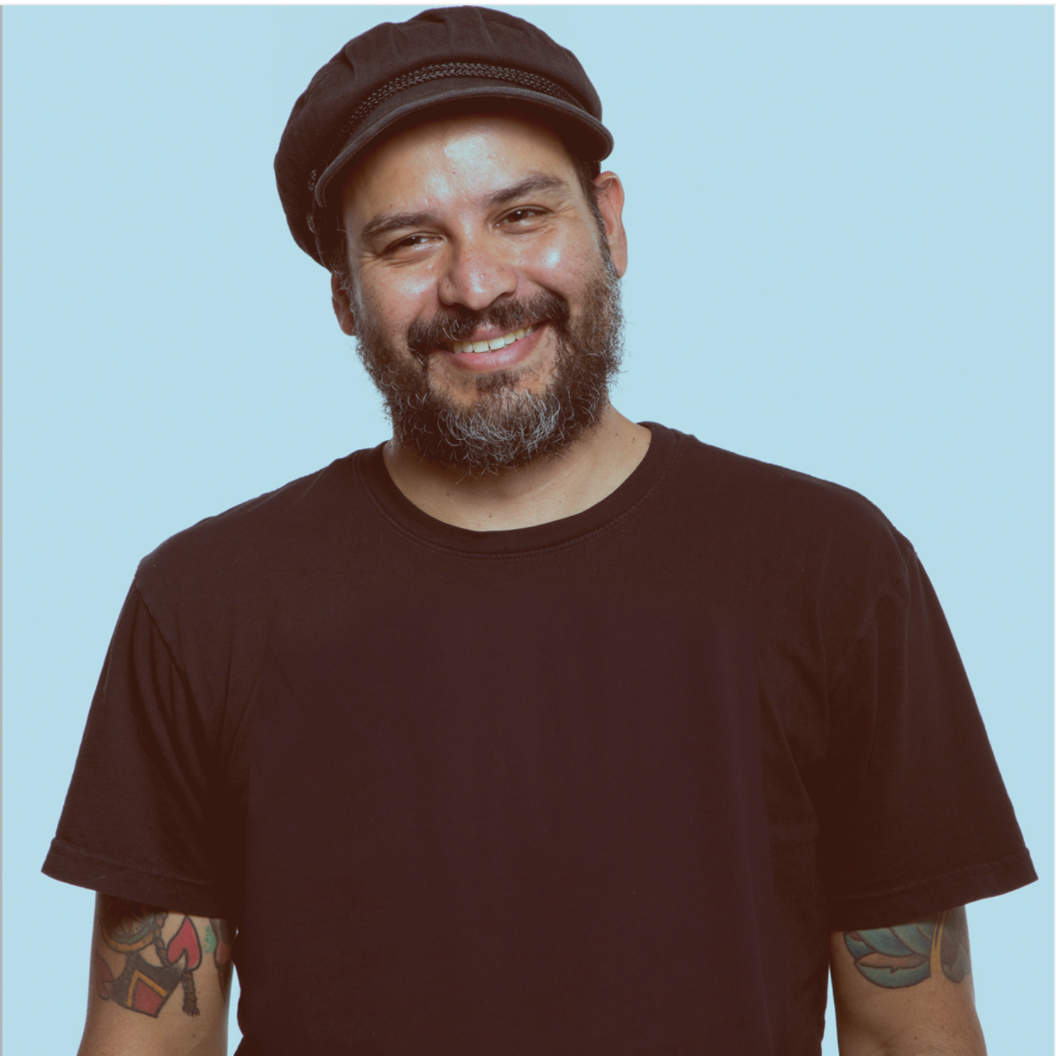 Man with a beard, wearing a cap and black shirt, smiling. Arms have tattoos, blue background.