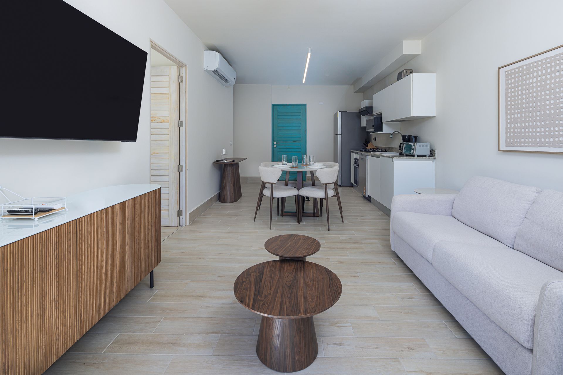 Sala de estar con sofá blanco, mueble de madera y mesas de centro redondas. Al fondo, el comedor y la cocina.