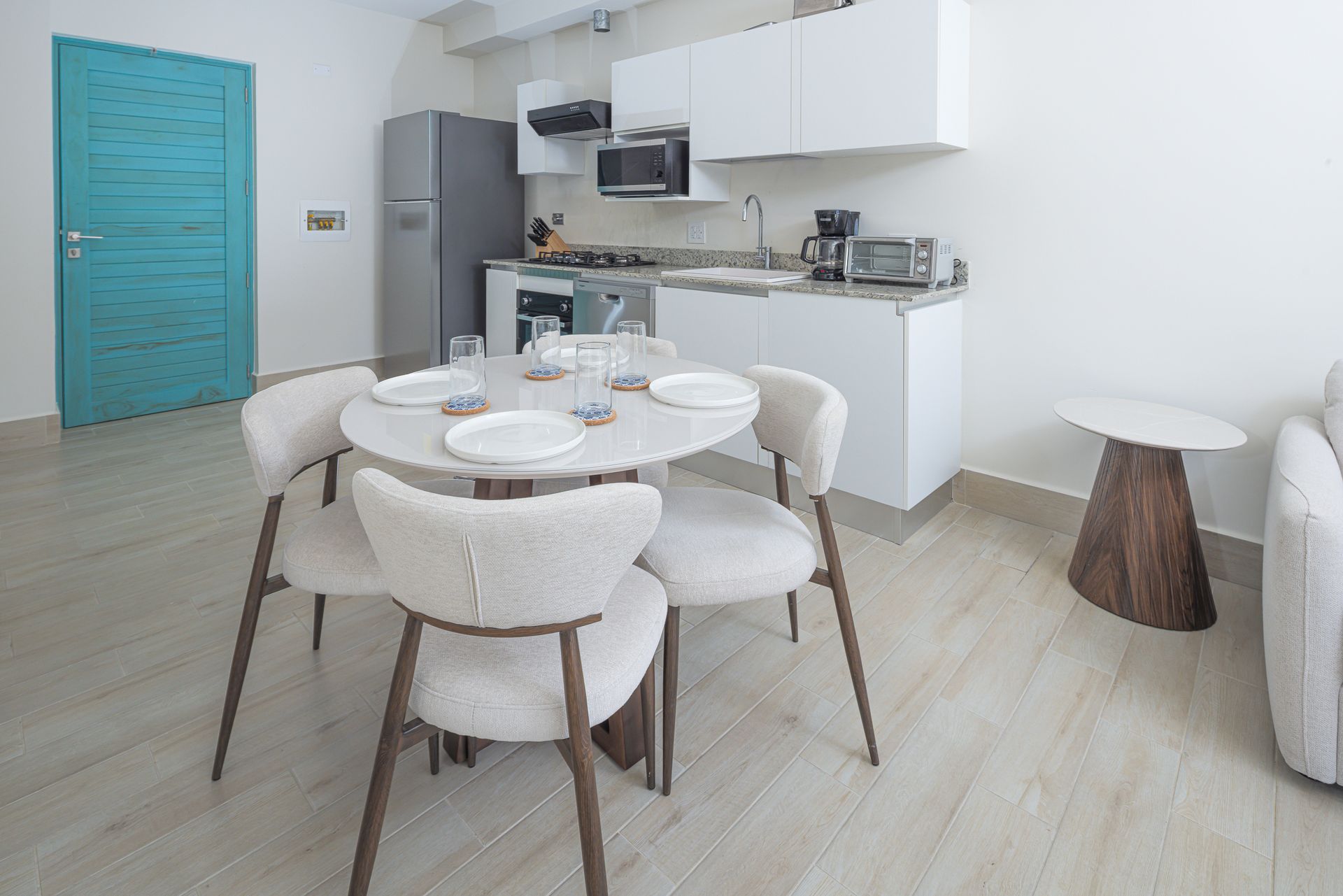 Una pequeña y moderna cocina y comedor con mesa redonda, sillas y muebles blancos. Puerta turquesa.