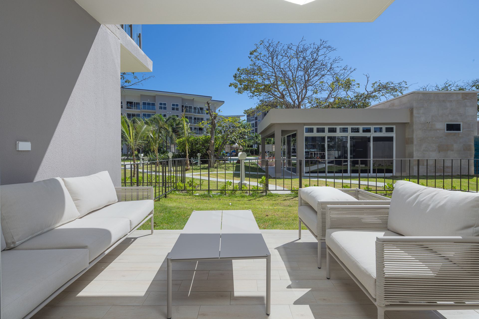 Patio con muebles blancos, paredes grises y un jardín con césped bajo un cielo azul.