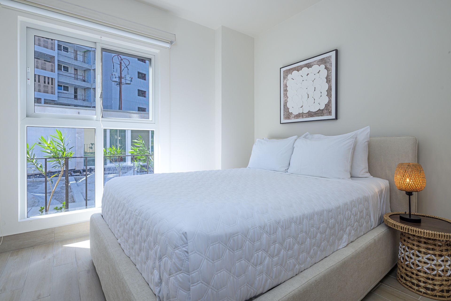 Dormitorio luminoso con ropa de cama blanca, pequeña mesa redonda con lámpara y gran ventanal.