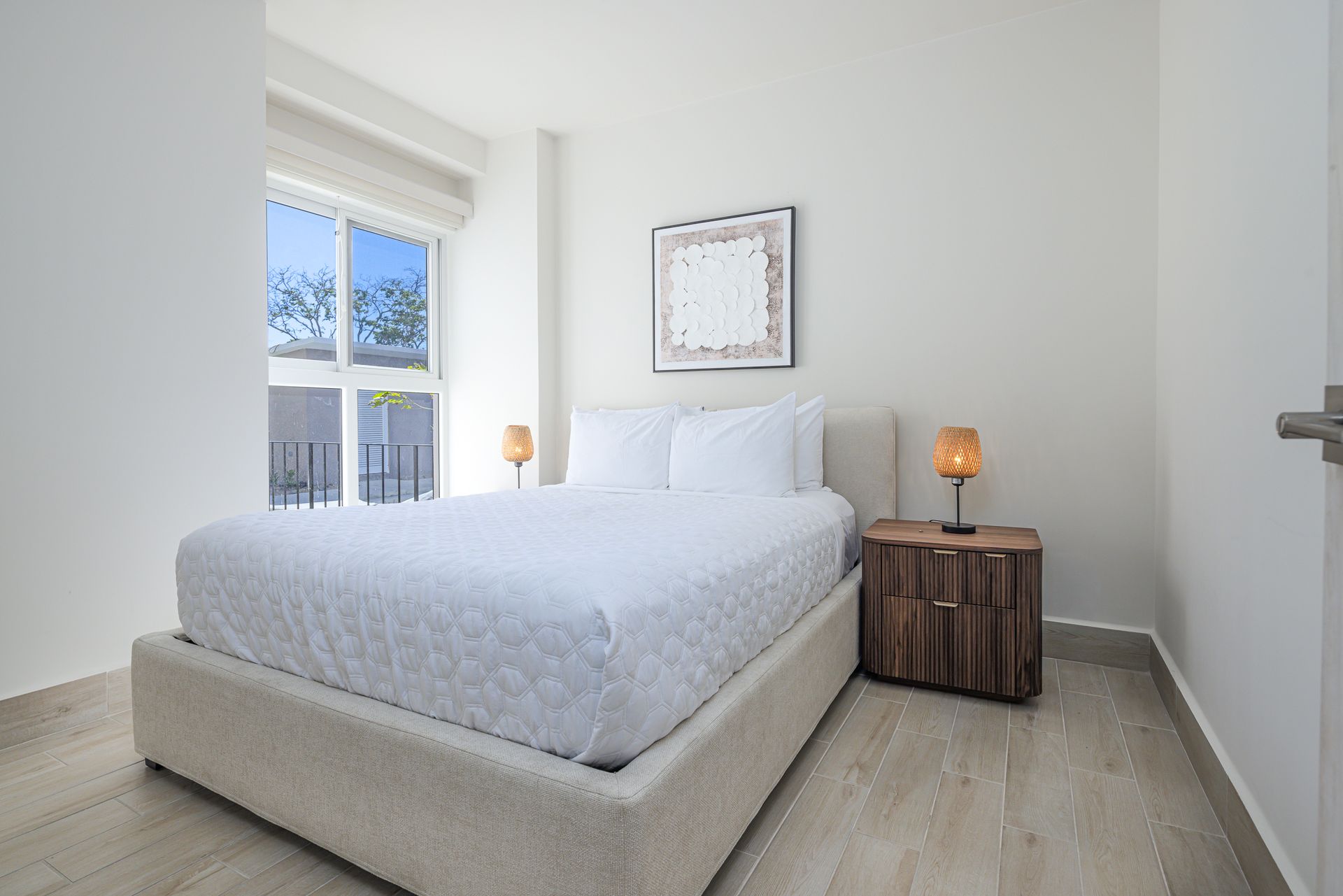 Dormitorio con cama, mesita de noche y ventana. Paredes y ropa de cama blancas.