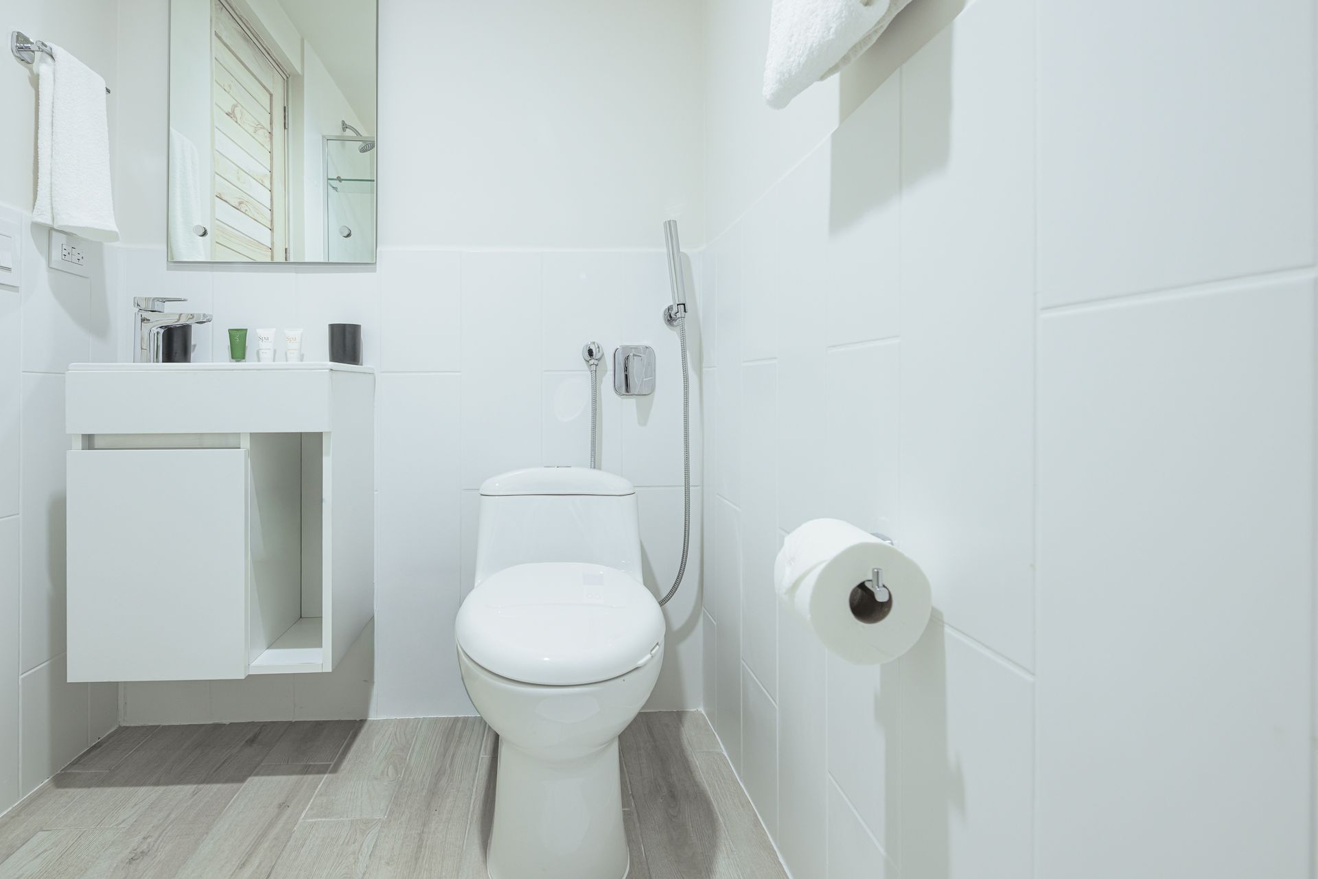 Baño blanco con inodoro, lavabo, espejo y rollo de papel higiénico.