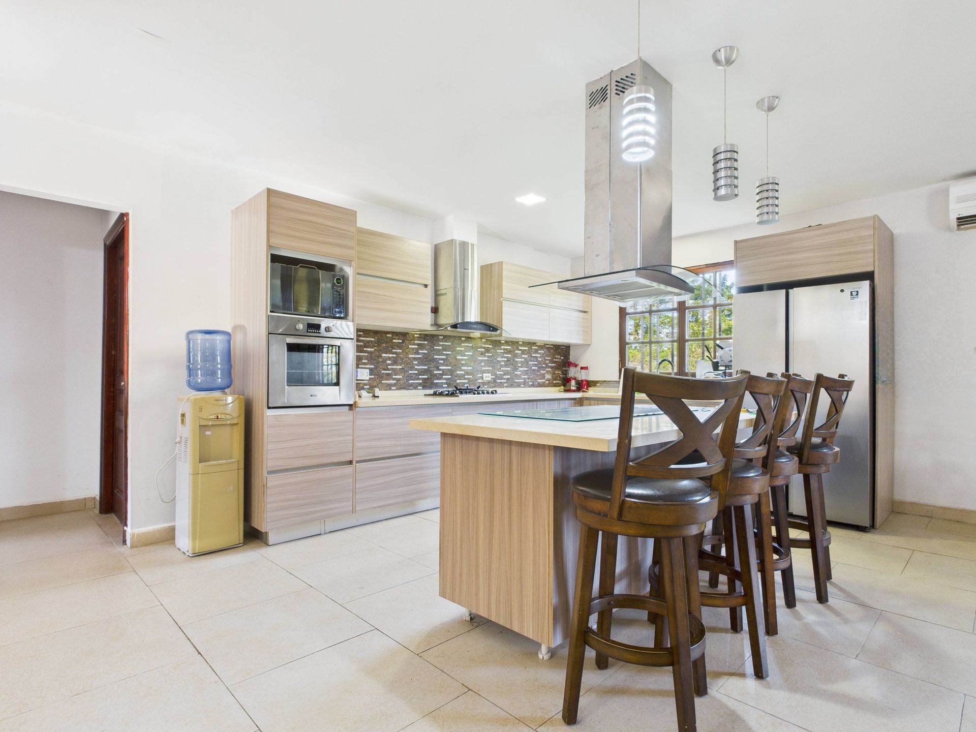 Cocina moderna con gabinetes de madera clara, isla con taburetes y electrodomésticos de acero inoxidable.