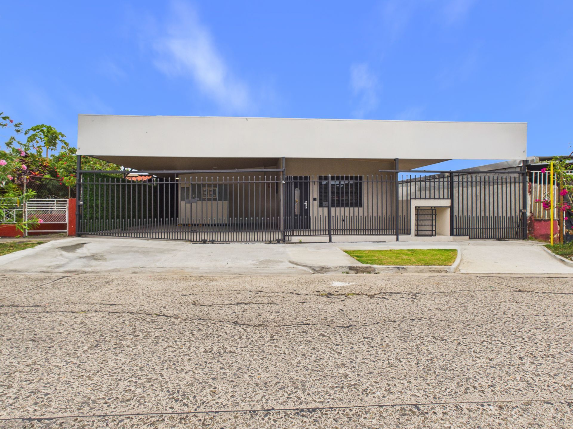 Casa de una sola planta con techo plano, patio delantero cercado y entrada central, vista desde una calle de grava bajo un cielo azul.