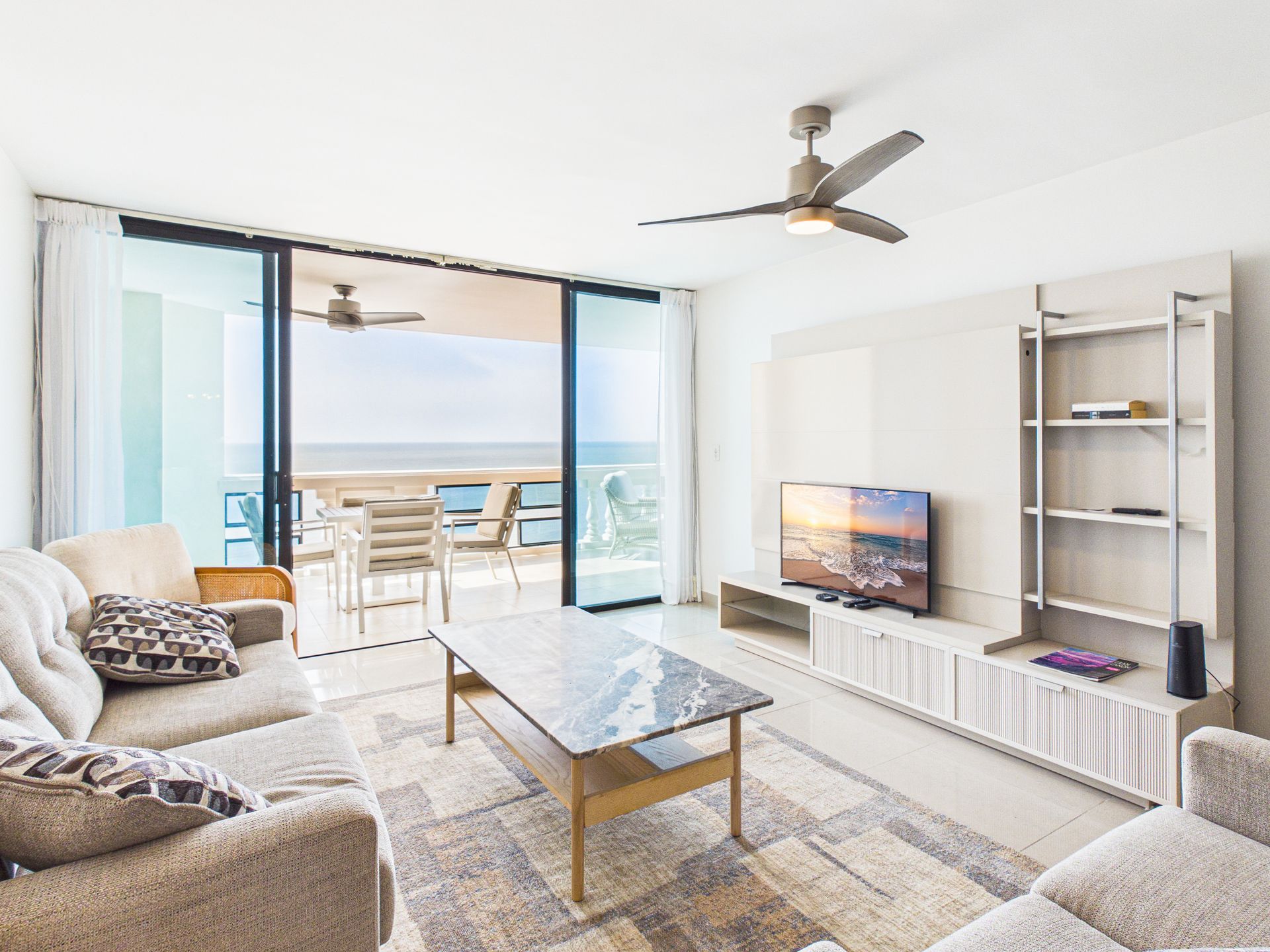 Sala de estar moderna con balcón con vista al mar, sofá beige, televisor, mesa de centro y ventilador de techo.coronado country club