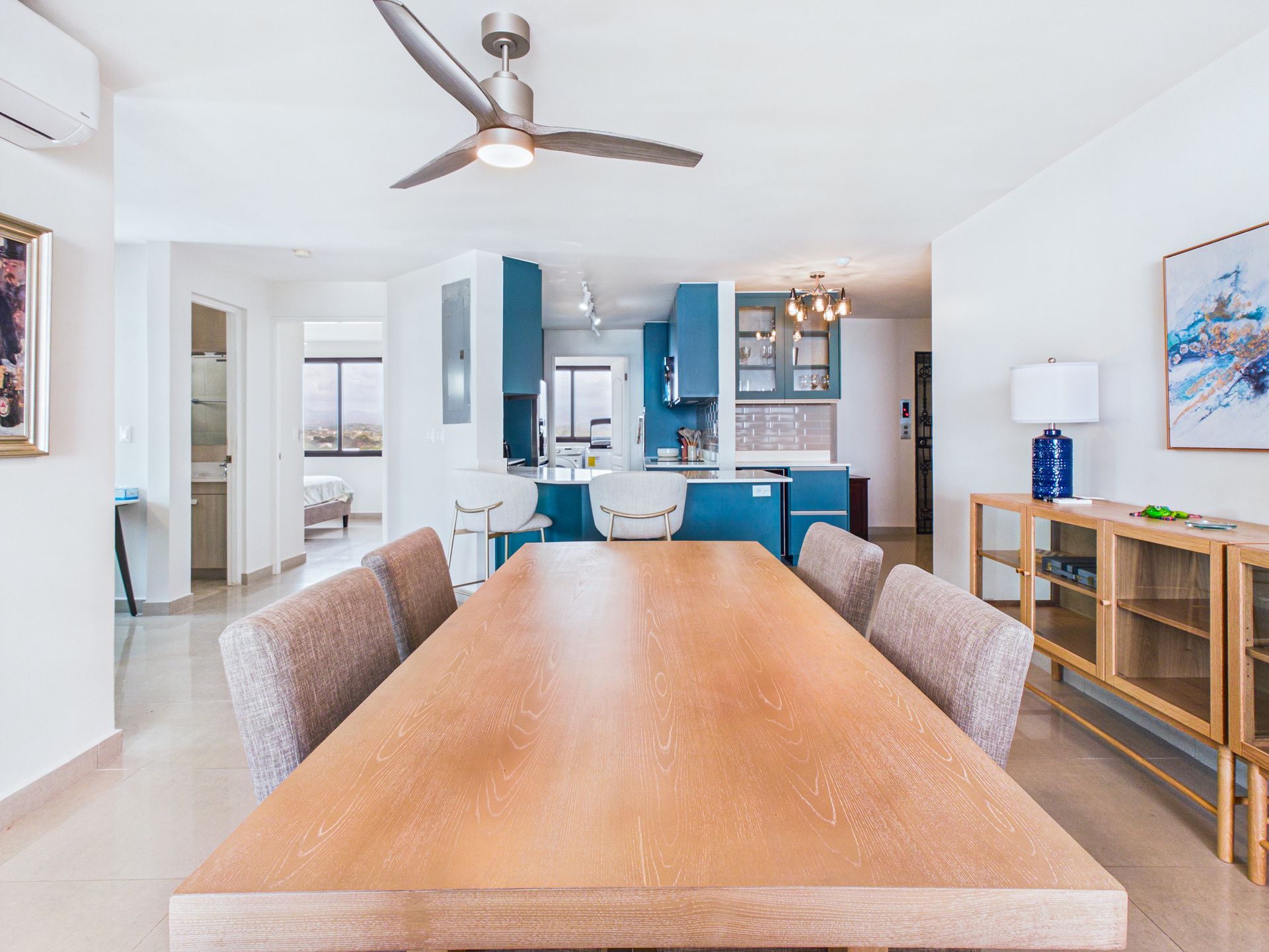 Comedor luminoso con una mesa larga de madera, sillas beige y una cocina azul al fondo.coronado country club