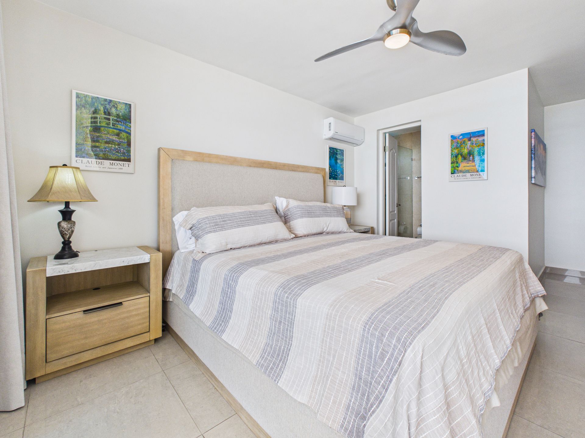 Dormitorio luminoso con cama de matrimonio, lámpara de mesita de noche, ventilador de techo y obras de arte en la pared, en una habitación blanca minimalista.coronado country club