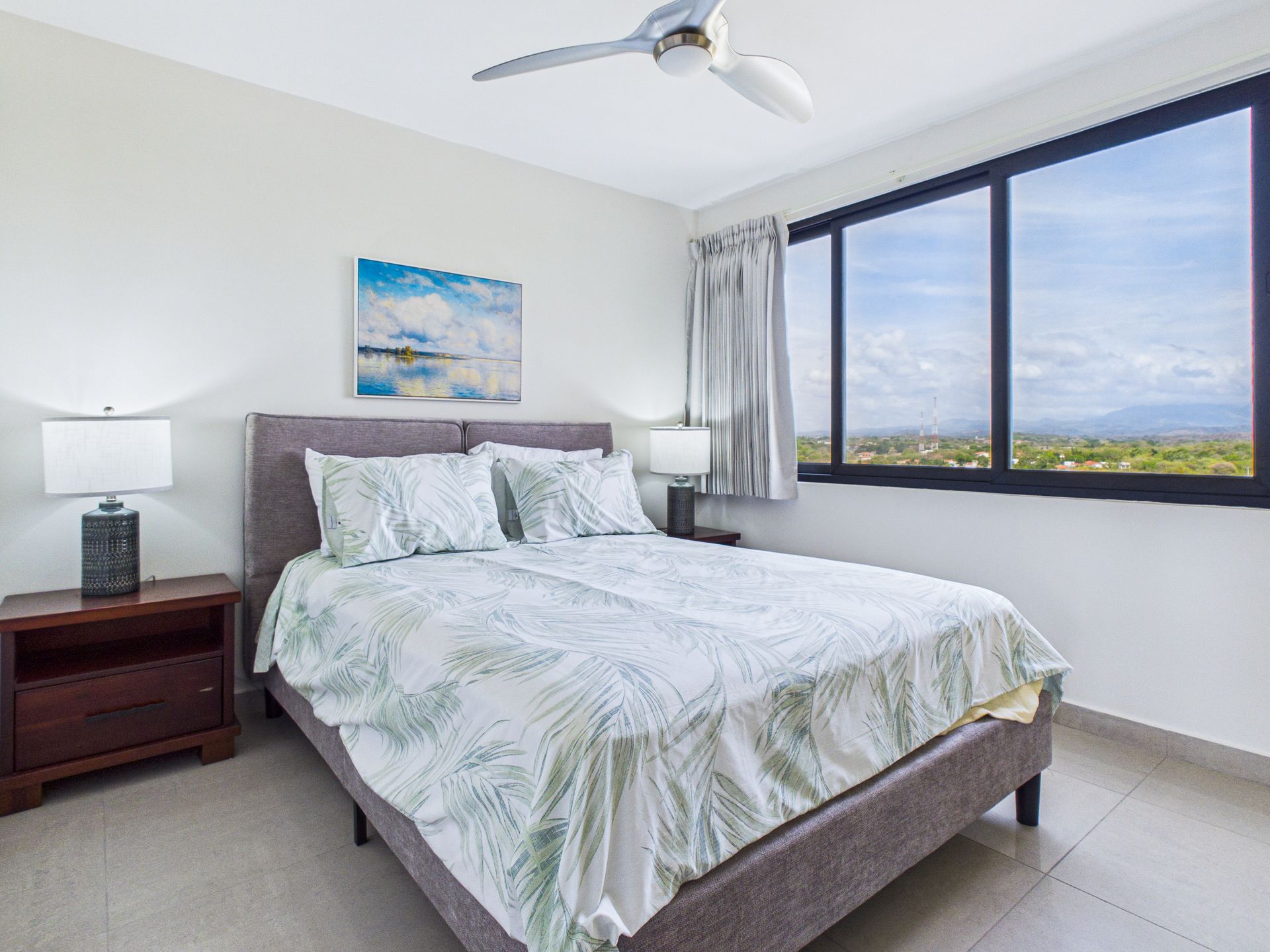 Habitación luminosa con una cama bien hecha, lámpara de mesilla de noche, cuadro en la pared y una gran ventana con vistas a la ciudad.coronado country club