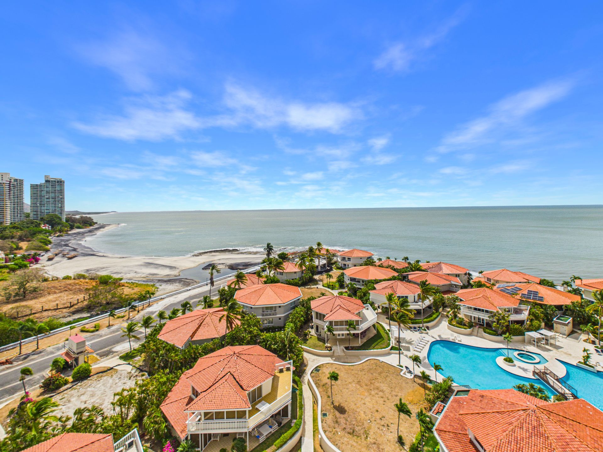 Complejo turístico costero con villas de techo rojo, piscina y vistas al océano bajo un cielo azul.coronado country club