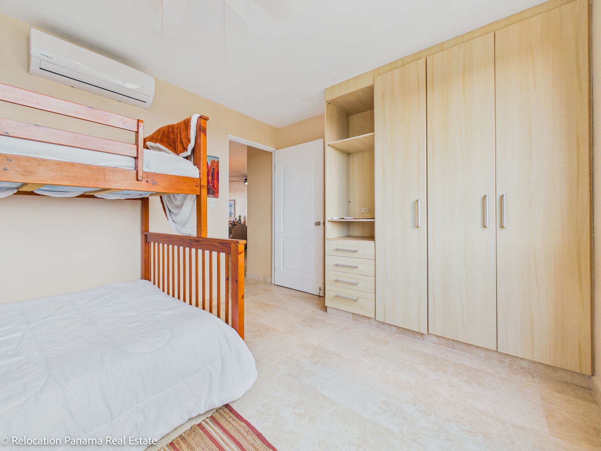 En una habitación luminosa con suelo de baldosas de color claro, un aire acondicionado blanco y una pared de armarios de madera clara, hay una litera.