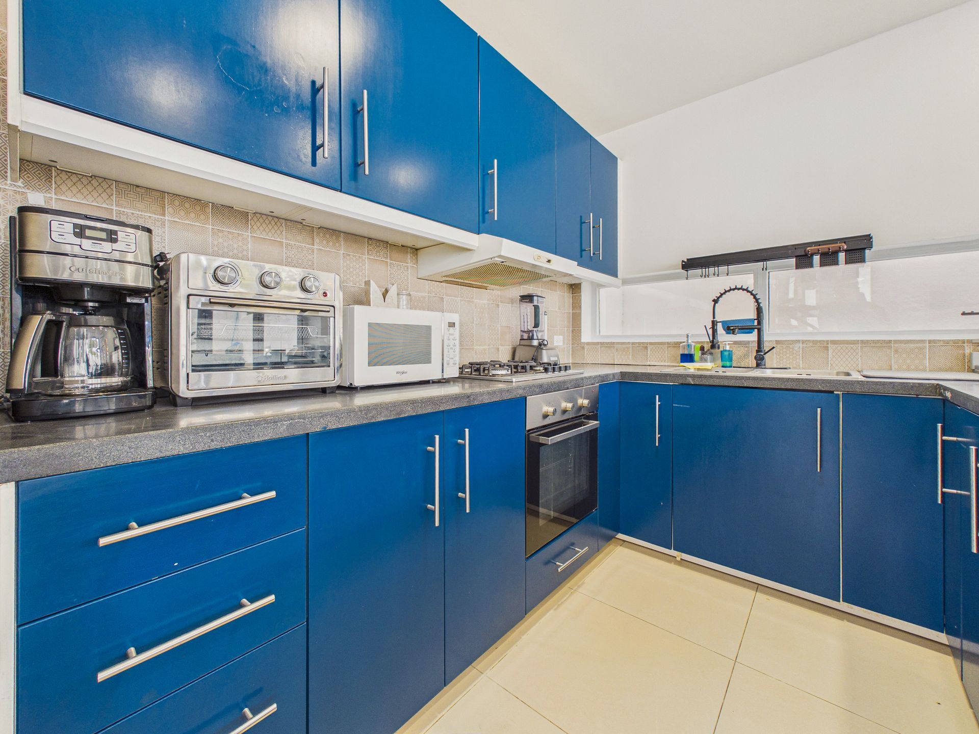 Cocina azul con armarios, electrodomésticos de encimera, fregadero y suelo de baldosas beige.